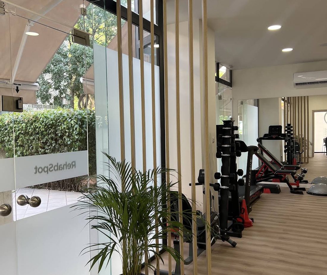 Η είσοδος του RehabSpot με ξύλινες περσίδες, φυτά και τον χώρο άσκησης στο βάθος